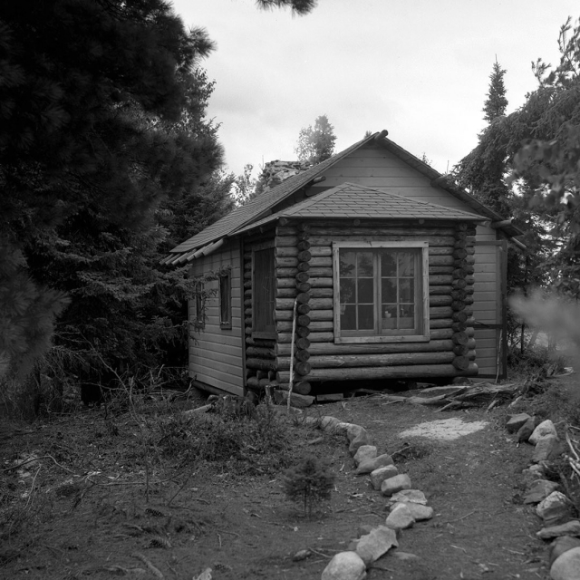 Black and white photograph of Jun Fujita's cabin on an island on Rainy Lake in Minnesota. Pine trees and other vegetation surround a cabin. A room addition bult of logs in foreground, with two large windows on its side. A stone-lined path leads to cabin 