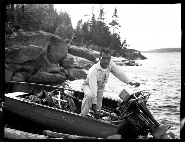 Historic circa 1931 photograph of Jun Fujita on a motorboat at the edge of a lake with boulders and outcrops and pine trees. 