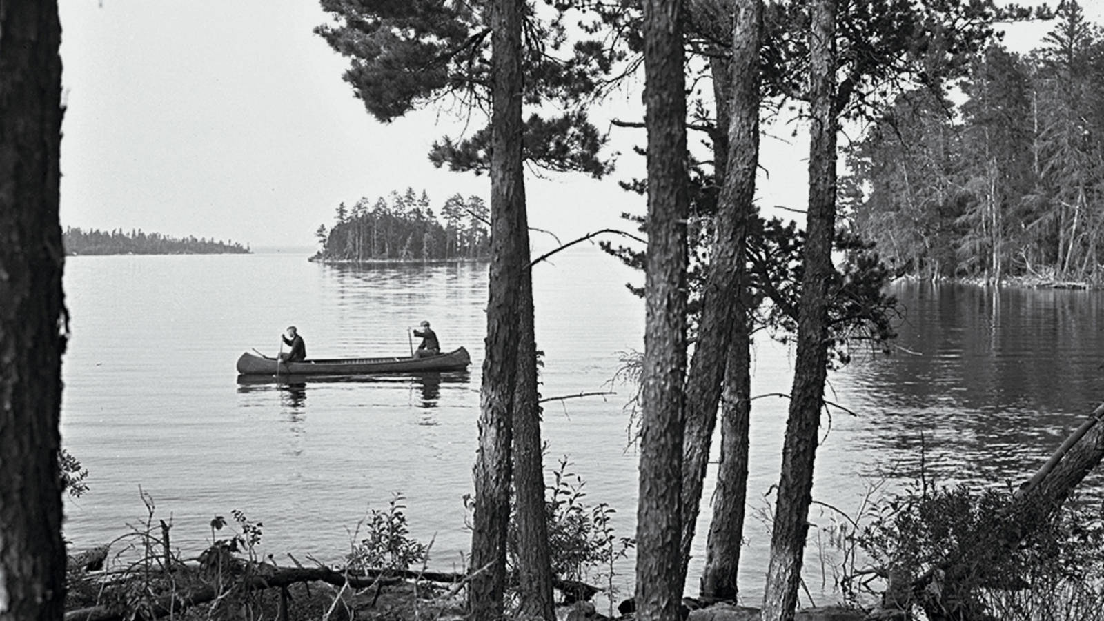Historic black and white photograph of two men in a canoe on a lake with rocky outcrops and islands covered in pines around them 