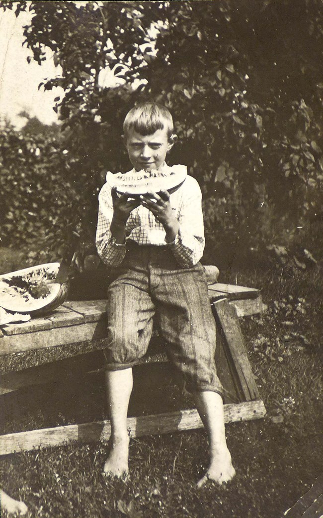 A 10-year-old boy sits on a crude wooden table with trousers on and a plaid shirt; eating a melon