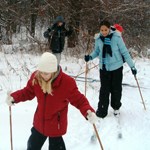 Three youths in cross-country skis, each using a set of poles to help propel them through snow. Leafless branches of a forest's edge make the background.