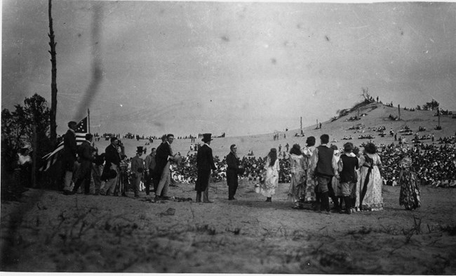 Historic photograph of 1917 Dunes Pageant; a line of costumed people stand atop a sandy stage area with a large sandy expanse beyond them. In the expanse are thousands of pageant attendees, dressed in typical 1910s clothing, watching the performance.