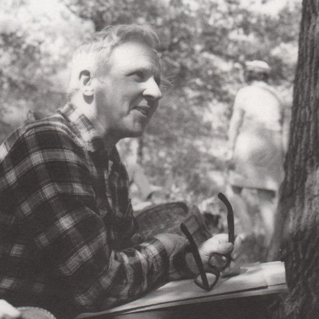 Black and white photograph from 1969 of a white-haired man holding a pair of glasses, wearing a dark plaid shirt, sitting next to a tree. A woman with a hat and gloves in background