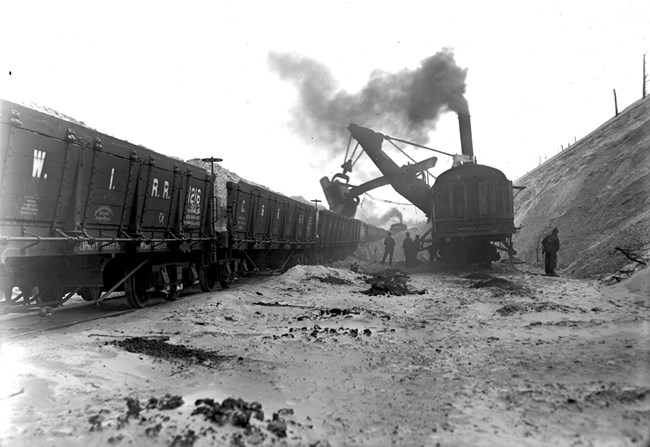 Historic black and white image of a steam shovel on a railrod track scooping sand from the Indana Dunes