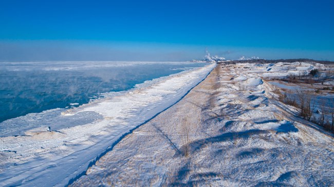 Aerial view of West Beach in winter. White shelf ice lines a snowy beach with snow-covered dunes. The blue of unfrozen Lake Michigan on left. Blue skies.