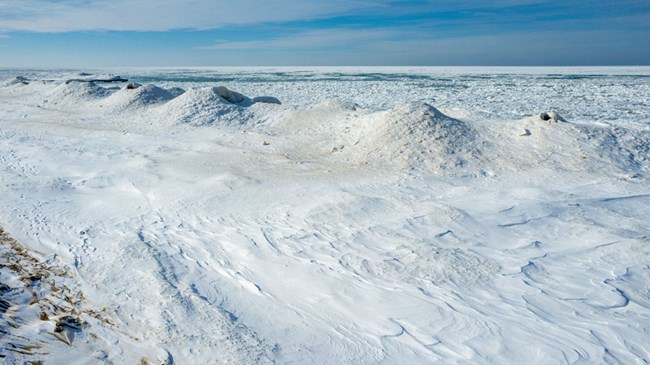 A row of white mounds diagonally extends along Lake Michigan's frozen shore. Blue skies with hazy clouds above.