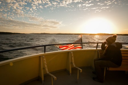 A person is sitting on a bench on the back of a large boat looking into the distance as the sun sets