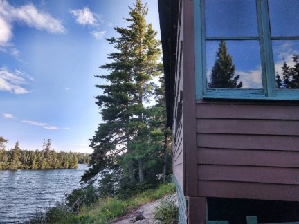 A view of the corner of a cabin on right with lake and trees on left