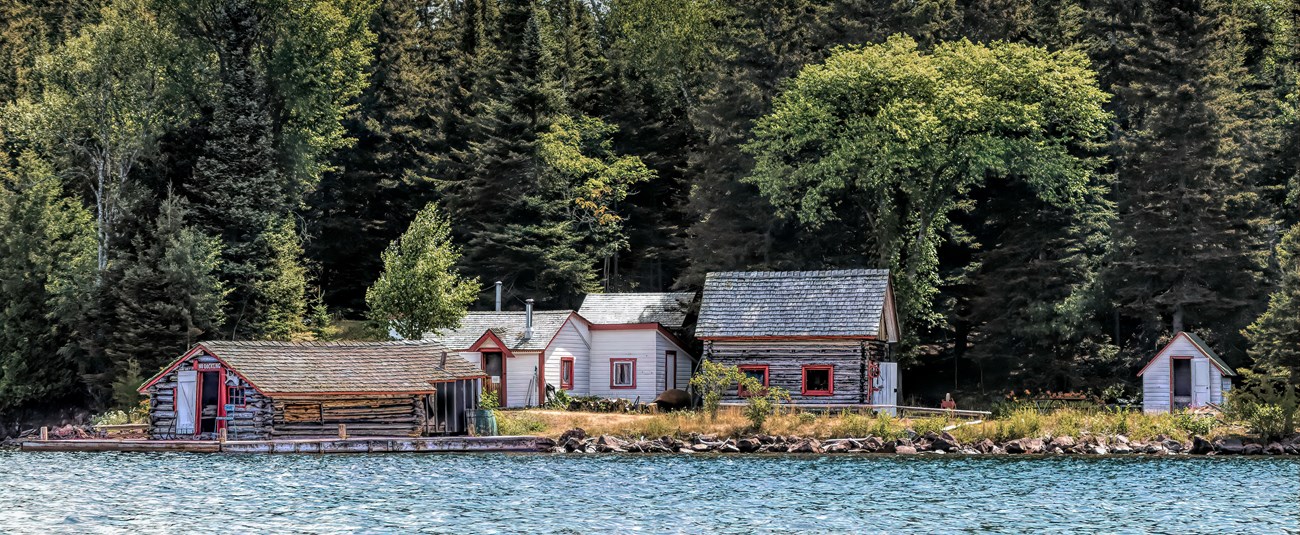 Multiple grey historic cabins and a lakeshore surrounded by forest.