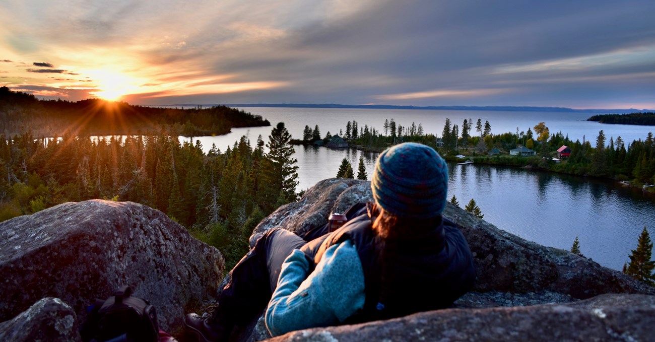 A person lays on a rock and view a sunset over an island and surrounding waters.