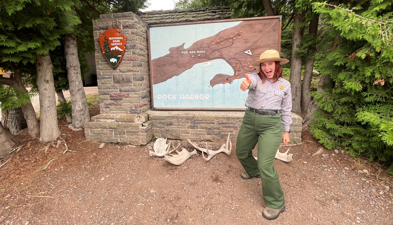 A park ranger with a big smile stands in front of a National Park Service sign with a big thumbs up.