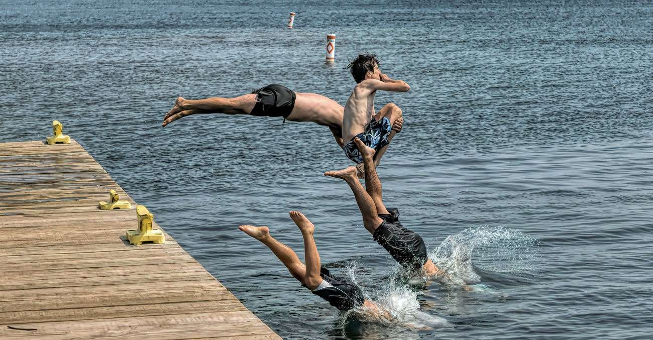 Three people jump and dive off a wooden dock into a lake.