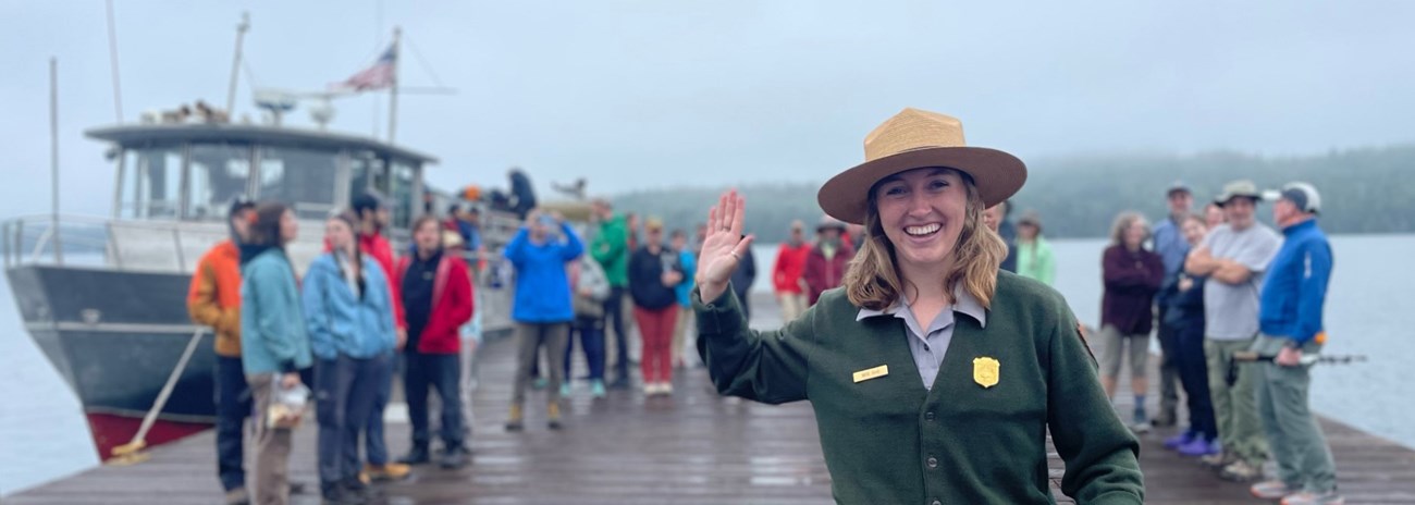 A smiling park ranger waves at the camera while a crowd stands behind her next to a ferry.