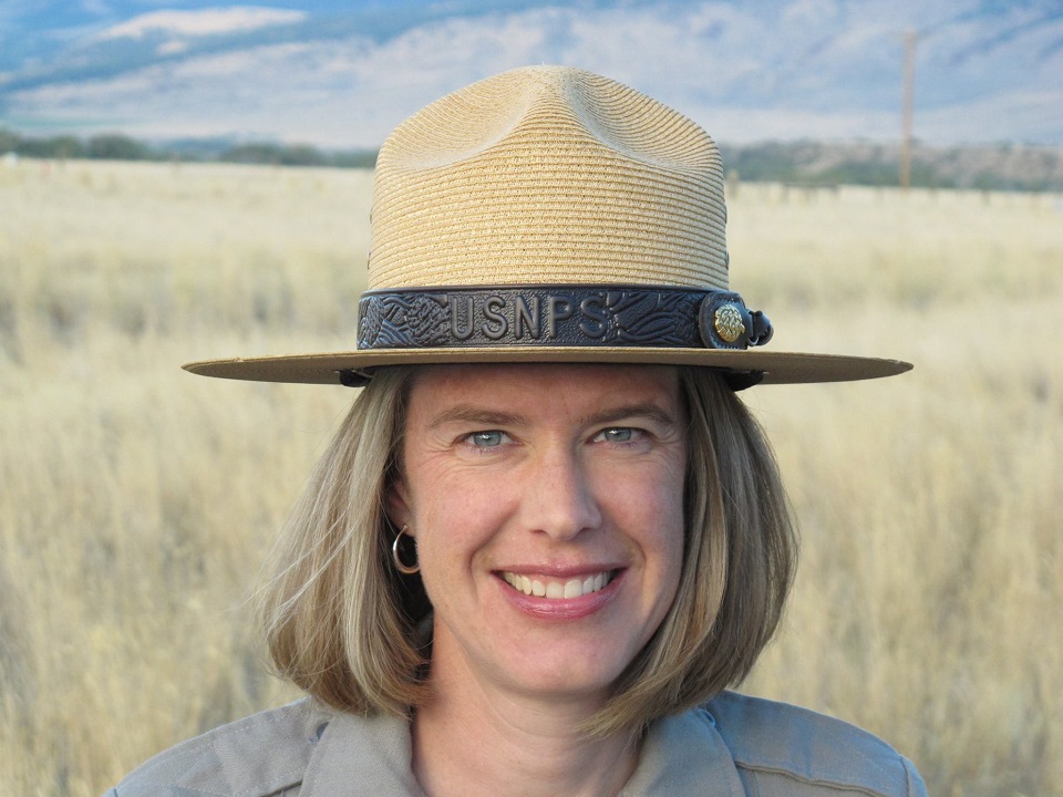 uniformed NPS employee poses for picture, field of grain and mountains in the background on a blue sky day