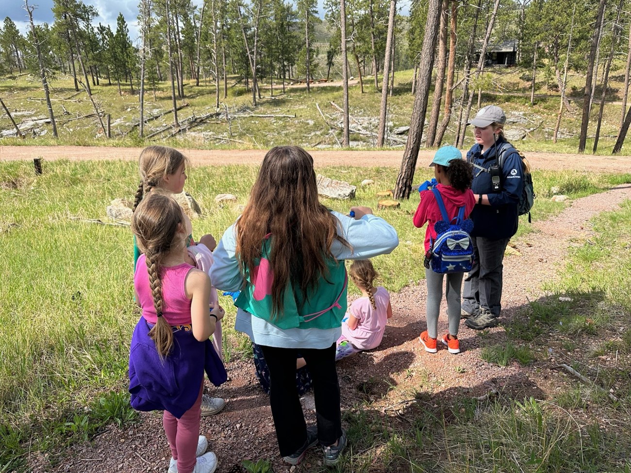 A ranger in blue stands with several youth on a trail through a pine forest.