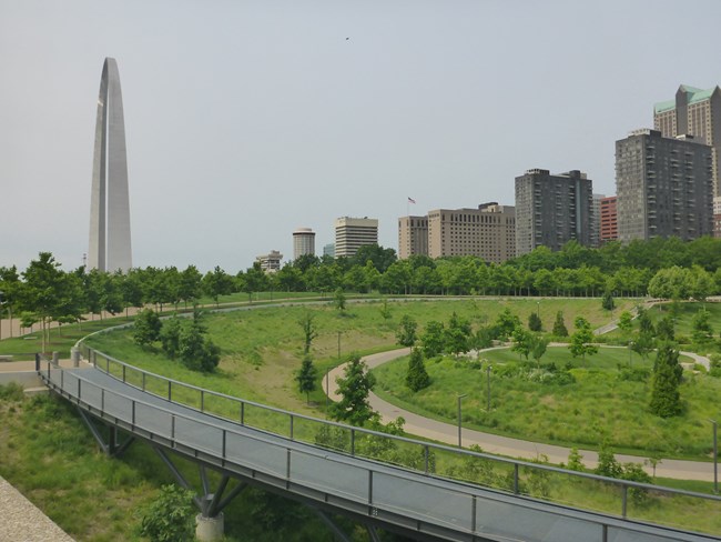 A green circular garden area with paved paths around it. A giant steel arch is in the background.