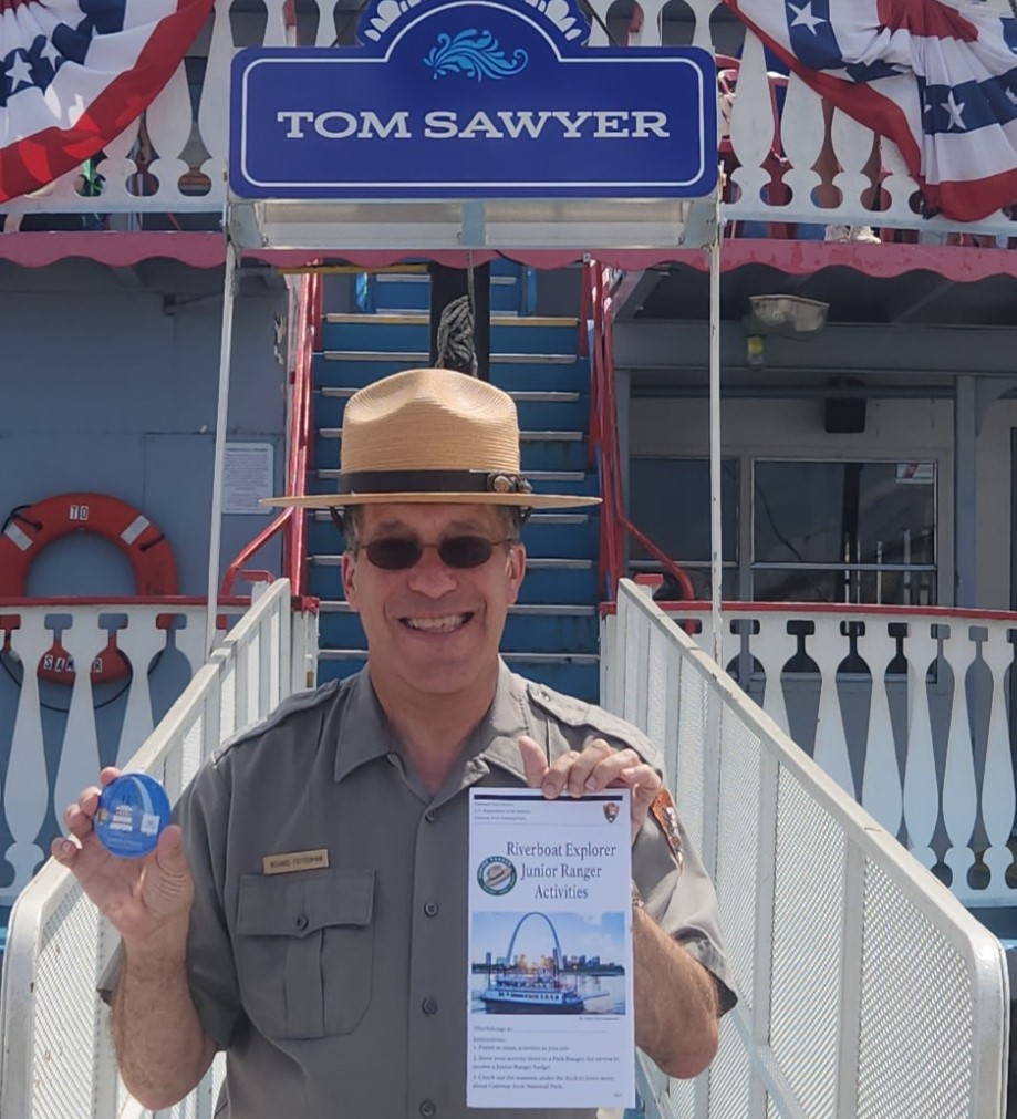 Male park ranger standing in front of the Tom Sawyer Riverboat. He is holding the Junior Ranger Riverboat booklet and the button you get when you complete the booklet.