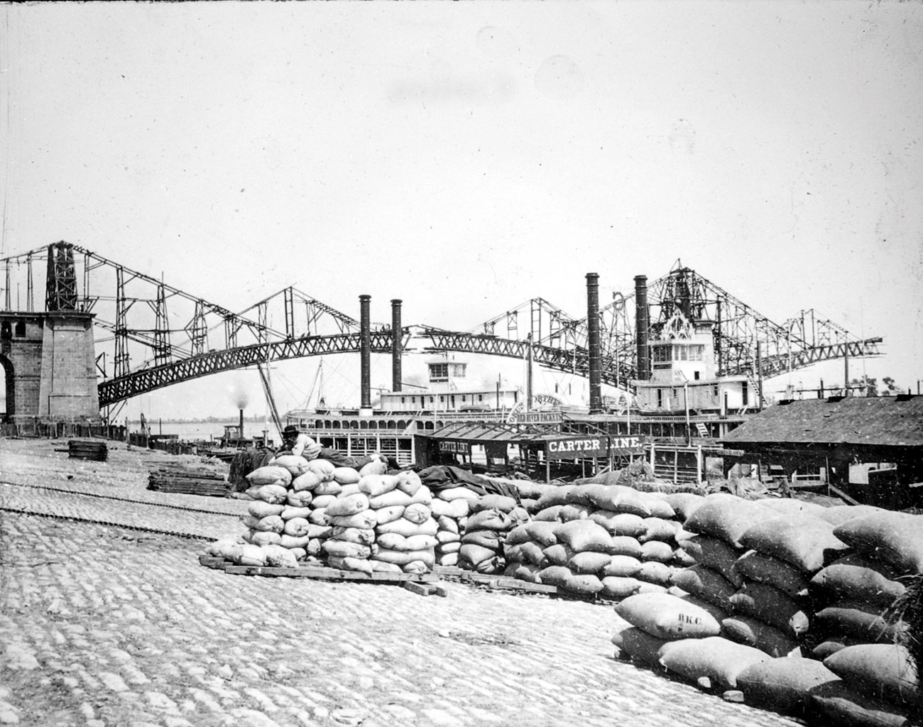 the steel frame of the bridge under construction in the background with steamboats and the cobblestone levee in the foreground