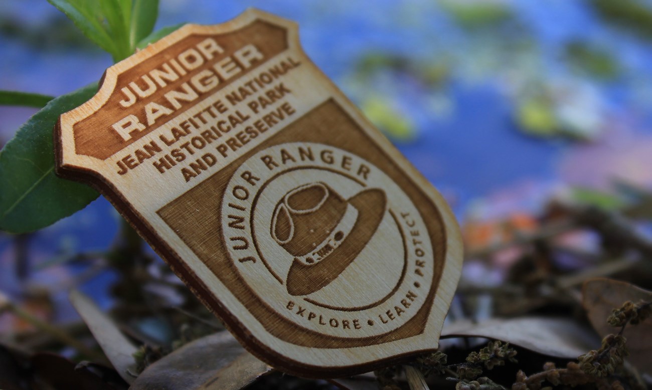 A wooden badge that says "Junior Ranger Jean Lafitte National Historical Park and Preserve" with a hat logo. Water is in the background.