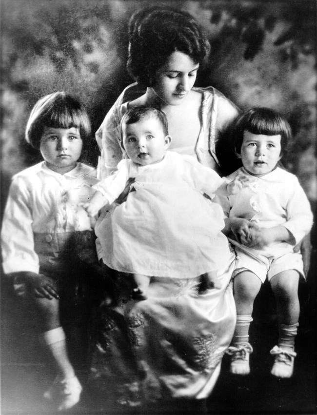 A black and white photo of a young woman in a formal gown sitting with a baby on her lap, and a small boy on each side. The two boys wear blunt haircuts, dress shirts, shorts, socks and shoes, while the baby wears a simple dress.