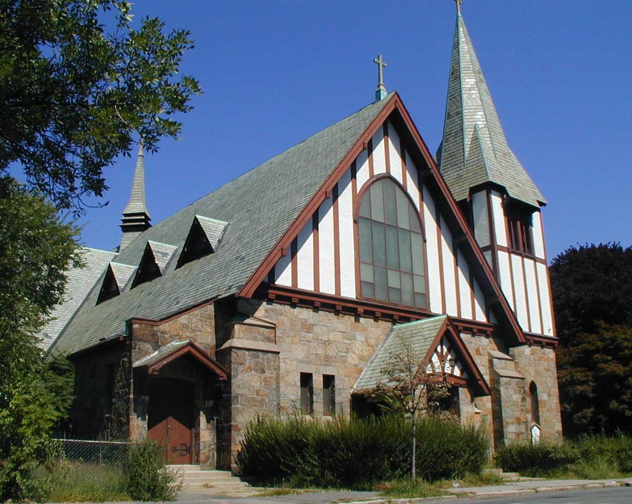 A photo of a church with stone walls, a white wooden gable and steeple with maroon trim, a multi paneled glass window, a maroon door, shrubbery in front and trees in the background.