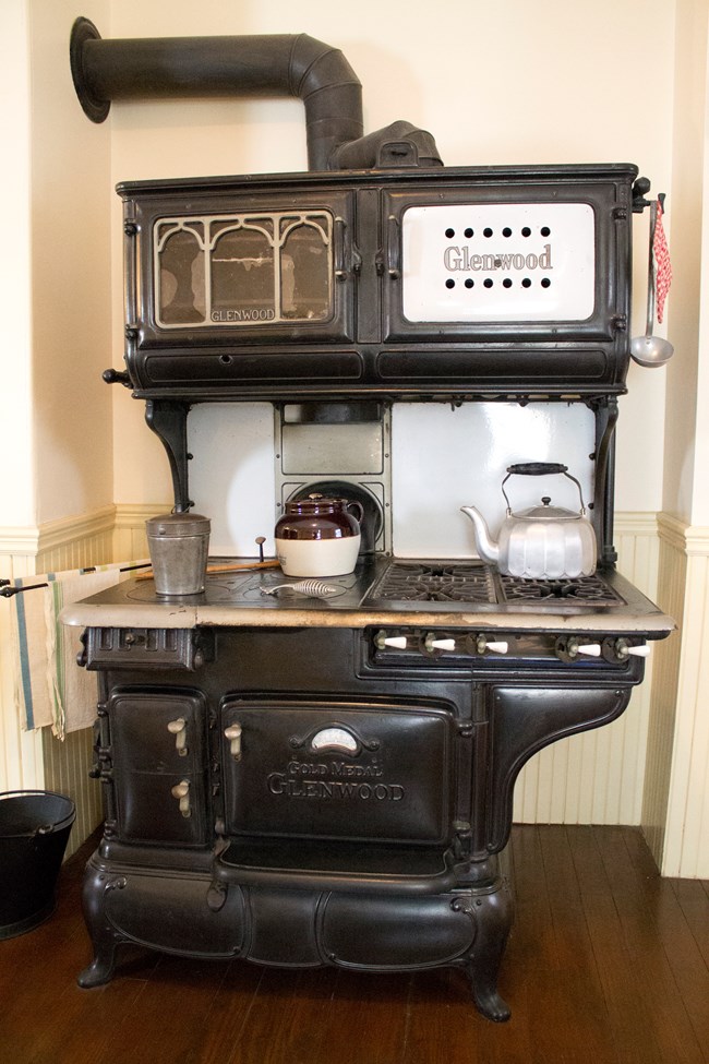 A photo of a large black stove, containing an oven, several burners, and compartments over the stovetop, whose chimney pipe connects to the wall. On the stove sit a bucket, a beanpot, and a tea kettle. A ladle hangs from a hook on the side of the stove.