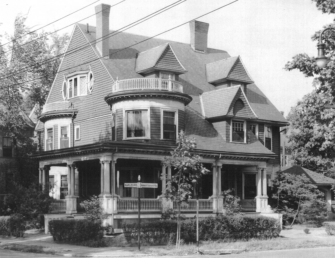 A black and white photo of a three story Victorian house with three gables, a bay window and rounded corner window with balconies, and a wraparound porch on a street corner.