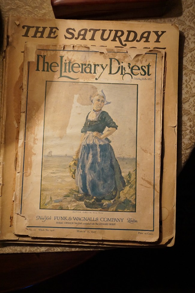 A photo of two frayed magazines. One partly reads ‘The Saturday.’ The other, dated ‘March 31 1917’ reads ‘The Literary Digest’ in large, swooping font, and shows a painting of a young woman in a blue dress and clogs standing by the seashore.