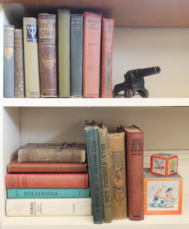 A photo of a white bookcase with two shelves, holding colorful, worn children’s books, toy blocks, and a metal toy cannon.