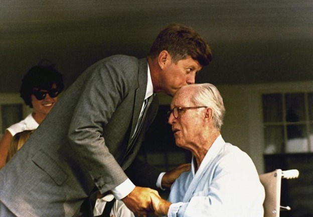 JFK kisses his father's forehead. He holds his father's hand with his right, his father's shoulder with his left. A sister smiles in the background.