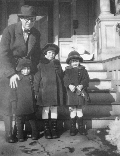 A man in a coat and hat stands with three small girls in front of the steps to a house. The girls wear matching winter boots, coats, and hats.