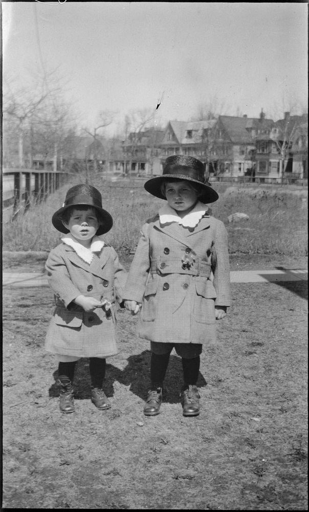 A black and white photo of two small boys in matching coats and hats holding hands, in front of empty, grassy lots, with a row of houses behind them.