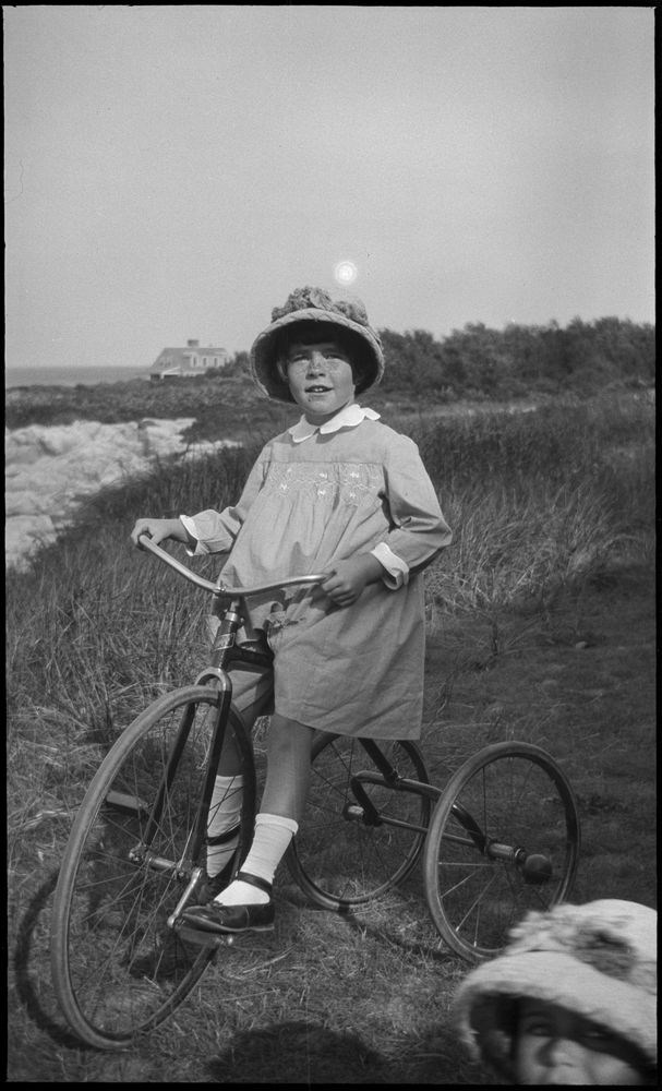 A black and white photo of a smiling young girl riding a tricycle on grass by the seashore. She has short hair and freckles, and wears a hat, dress, and black shoes. A house sits on a rocky shore in the background. A girl in a hat is in front.