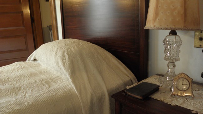 A photo of a wooden nightstand next to a twin bed covered with an embroidered white blanket and pillowcase. A small leather bound bible sits on the nightstand, next to a glass lamp and a clock encased in silver, whose face reads three o’clock.