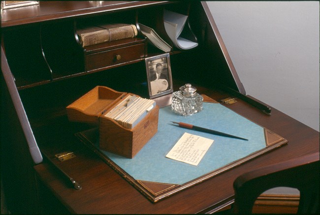A photo of a wooden desk holding books, a framed photograph of a man holding a baby, a writing pad, a glass paperweight, a pen, and a small box of paper cards, one of which is on the desk.
