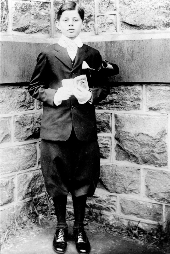 A black and white photo of a boy in a white collar and tie, dark blazer, breeches, and dress shoes standing against a stone wall, holding a small book with an image of Jesus Christ on the cover.