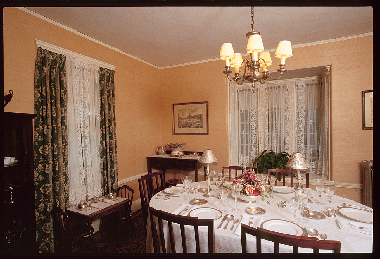 A photo of a dining table, set with china, glasses, and silver utensils. By a lace curtained window sits a low table with two chairs, set with silver porringers and utensils.