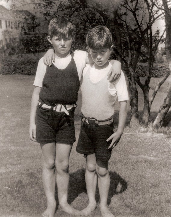 A black and white photo of two boys, one taller, standing with their arms around each other’s shoulders, wearing shorts and tee shirts under tank tops, barefoot on a grassy lawn. A large white house is in the background.