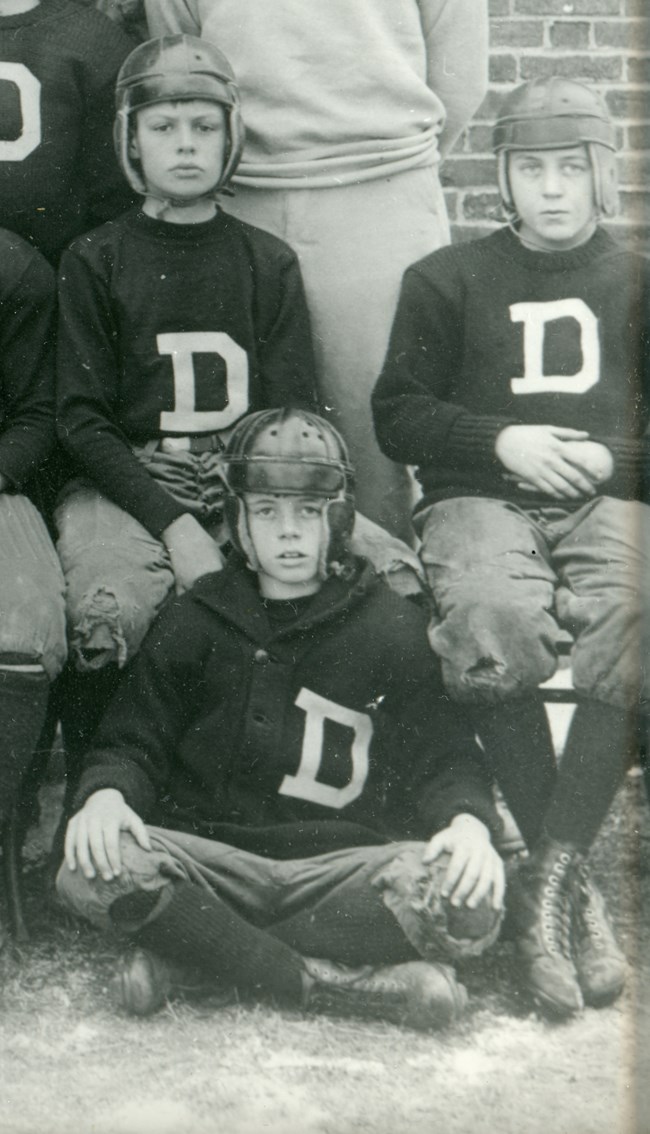 A black and white partial football team photo, with three boys visible in leather helmets, dark sweaters with a white letter “D” on the chest, trousers, knee socks, and boots with laces. Two sit on a bench, one sits on the grass in front.