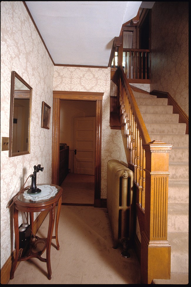 A photo of the home’s front hallway, with a staircase on the right. A black candlestick style telephone sits on a table in front of a mirror. The kitchen is partly visible in the background.