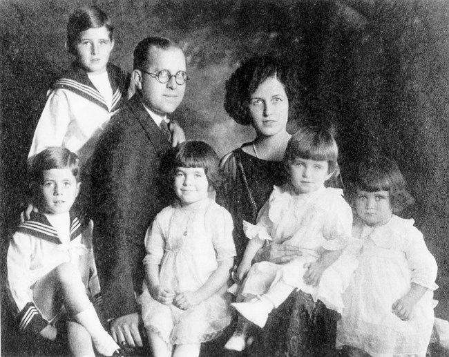 A black and white family photo. A man in a dark suit and glasses sits by a woman with short hair in a dark gown. Two boys in sailor suits stand and sit left of the man. Three girls in white dresses and bob haircuts sit on and next to their parents’ laps.
