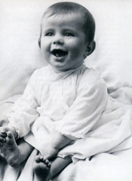 A black and white photo of a laughing baby in a white dress sitting on white fabric. The baby is barefoot, and is holding its right foot with its right hand. The baby has light eyes and a full head of downy hair.