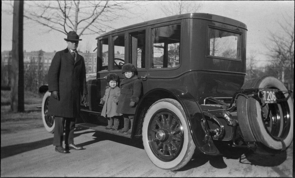 A black and white photo of a man in a coat and hat standing beside two small boys in matching coats and fur hats, who stand on the running board of an automobile, a building and trees in the distance.