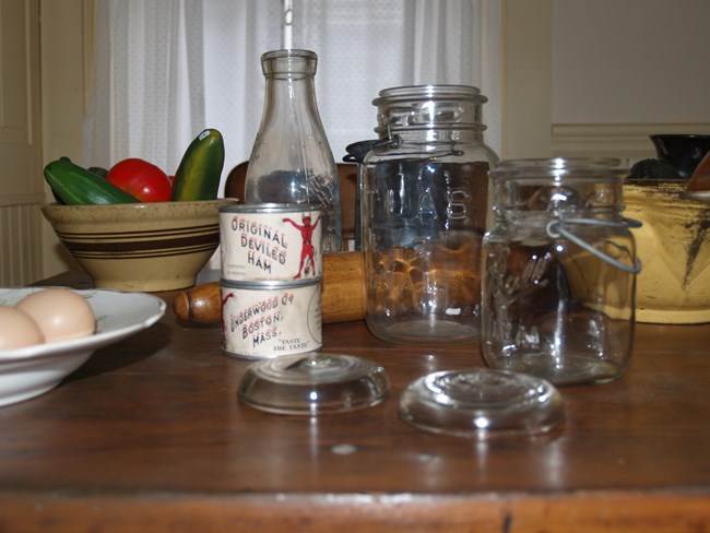 A photo of items on a wooden tabletop, including a dish of plastic eggs, a bowl of plastic vegetables, antique deviled ham cans, a glass milk bottle and mason jars, a wooden rolling pin, a pail, a metal stirring bowl, and a food chopper.
