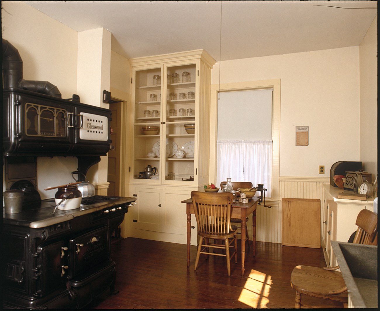 A photo of a small kitchen with off white walls. On the left is a black stove, on which sit a bucket, a beanpot, and a teakettle. In the back left corner is a cabinet, containing glass jars, ceramics, fine china, and other utensils.