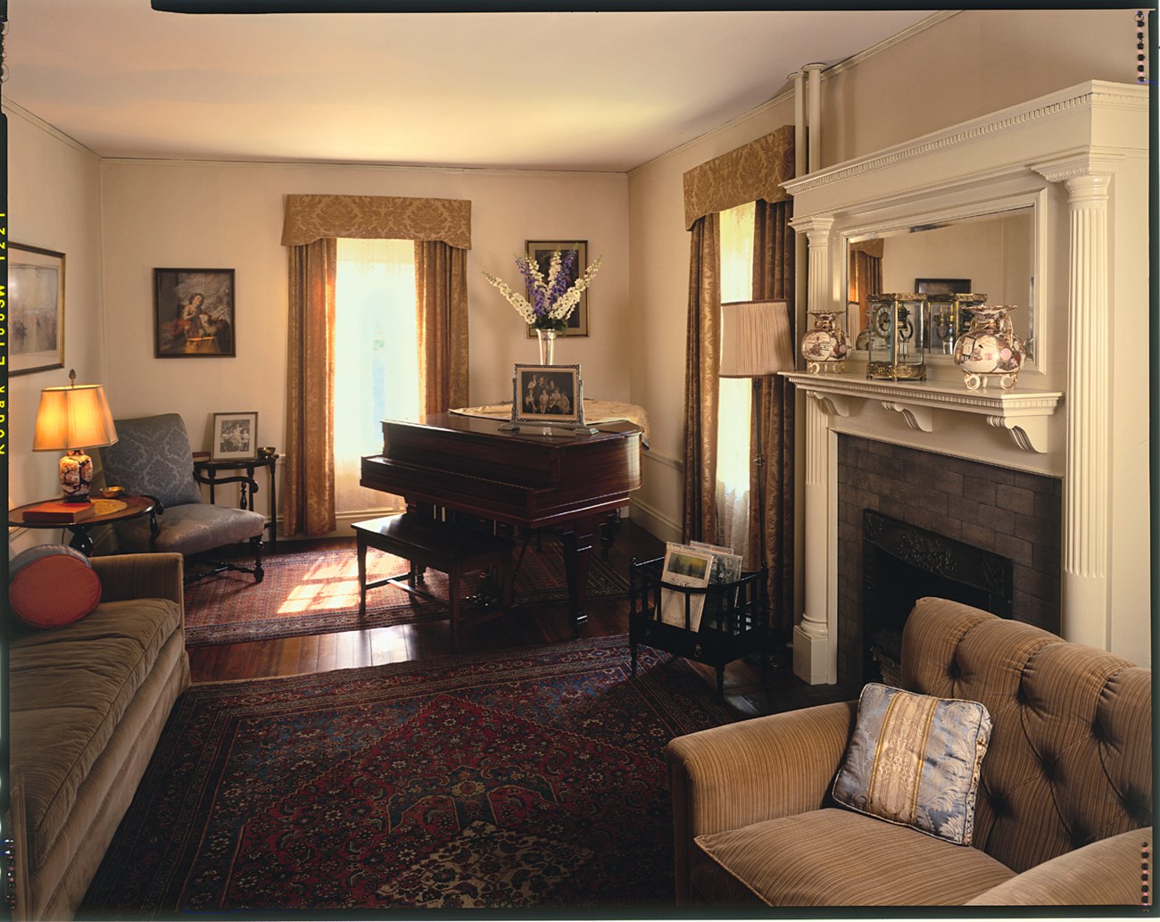 A photo of a sunlit room with a piano, fireplace, couch, armchairs, and lamps. Rugs cover the floor, paintings hang on the walls. Japanese vases and a clock sit on the fireplace mantle. By the piano are books in a bin, family photos, and a flower vase.