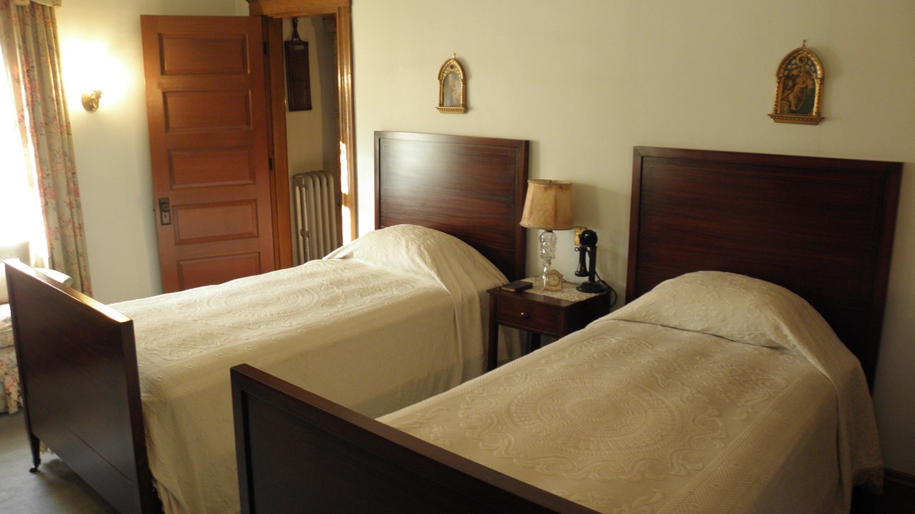 A photo of a room with two twin beds, a hallway door, and a window. On a nightstand between the beds is a lamp, a black candlestick telephone, a book, and a clock. Above each bed is a portrait of the Madonna and child.