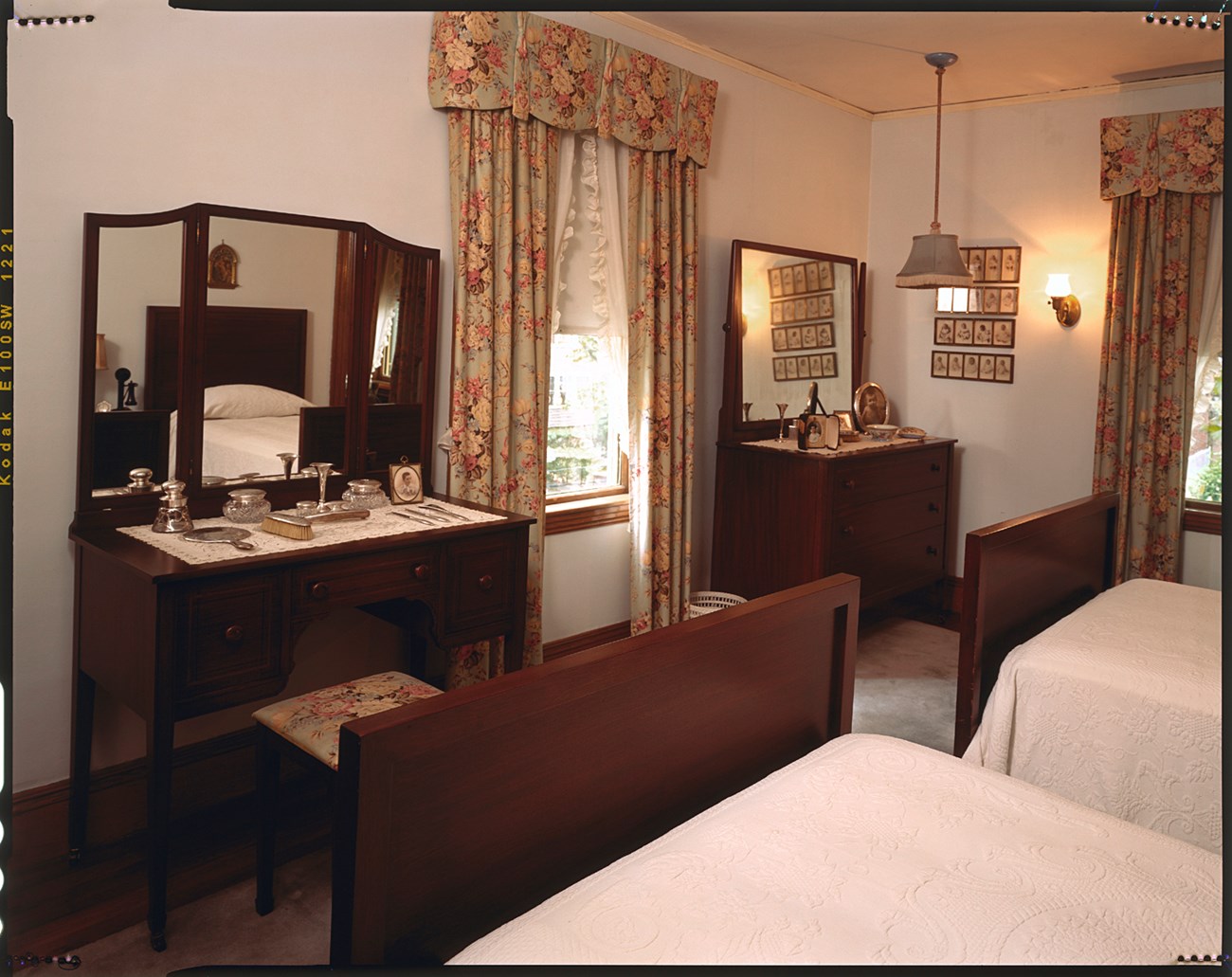 A photo of two dark wood vanities on each side of a window, across from two twin beds. On one vanity is a set of silver grooming tools, including a mirror and hairbrush, and a framed photograph. On the other are more photographs.