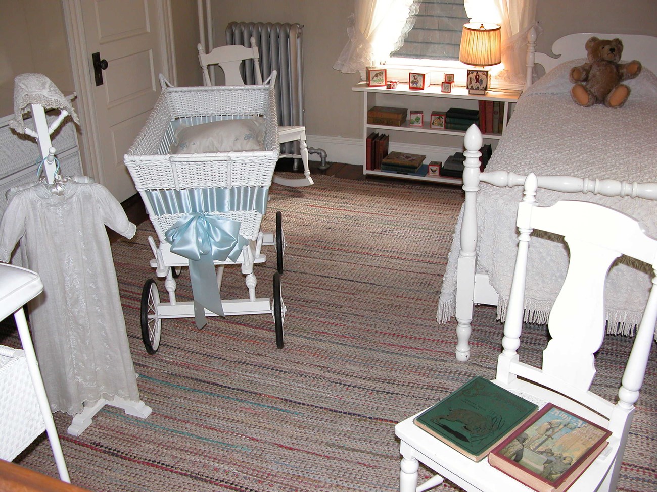 A photo of a nursery with white furniture: a clothes rack with a white gown and cap, a wheeled crib, rocking chair, a nightstand holding toy blocks, books, and a lamp, a bed with a teddy bear on it, and two books on a chair.