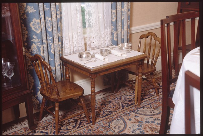 A photo of a small wooden table and two chairs, set with silver porringers, a bowl, spoons, napkin rings and cloth napkins. The table sits on a rug by a window, next to a china cabinet and behind a regular sized table.